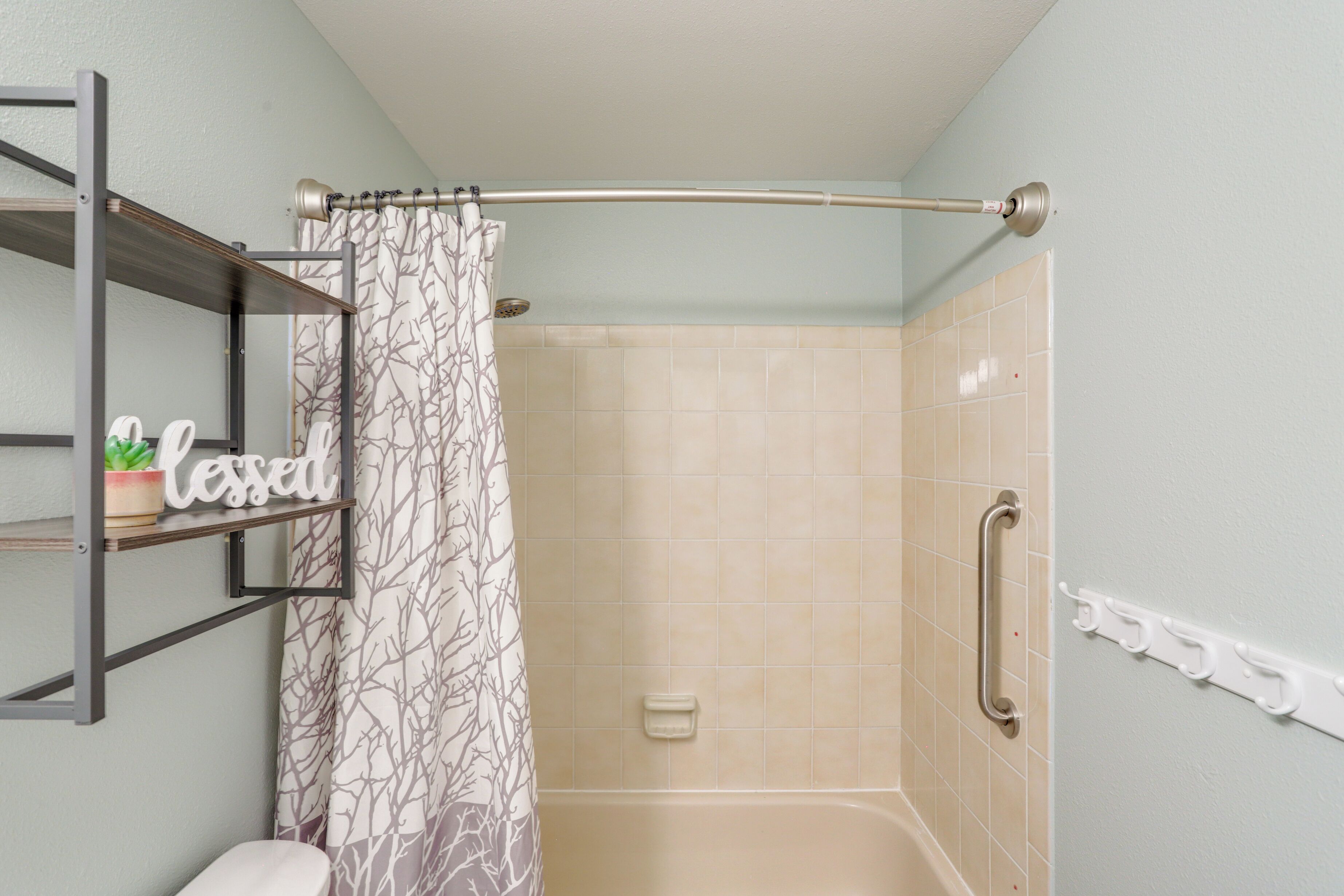 Zona de ducha con pared de azulejos color beige, cortina de baño con estampado de árboles, barra de apoyo y repisa con planta en maceta y un letrero que dice "lessen".