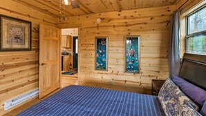 A wooden cabin interior with a bed, a ceiling fan, and framed artwork on the walls. - Cozy Mtn Cabin: Spacious Deck & Forest Views! (Murphy)