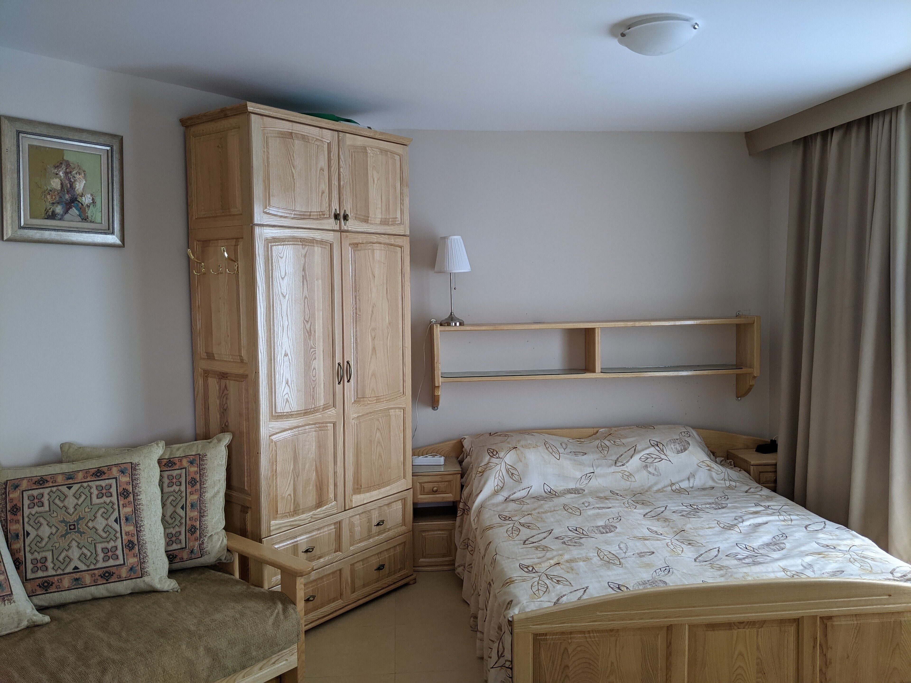 A bedroom with a wooden wardrobe, a bed with patterned bedding, a bedside table, a lamp, and a chair with cushions.