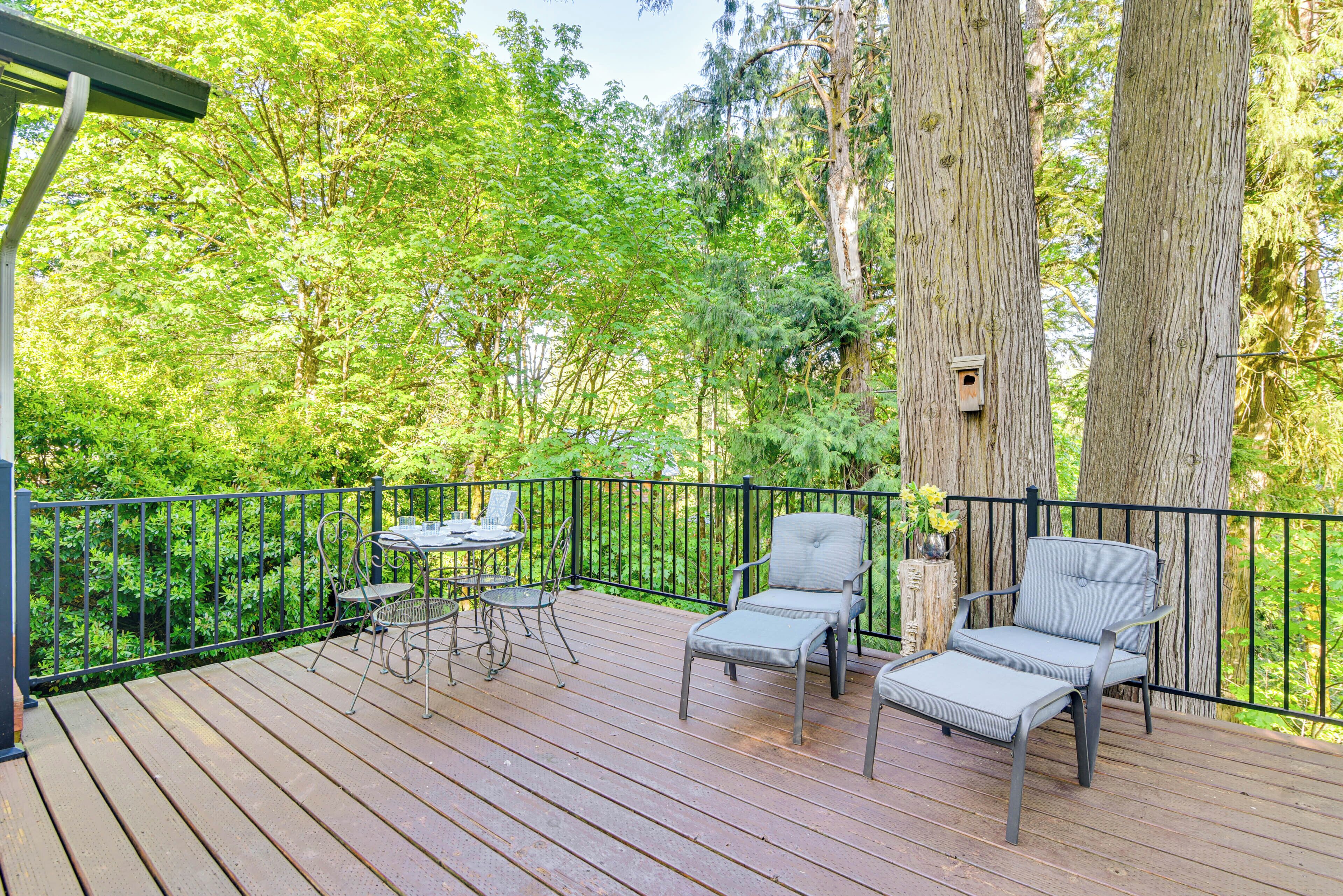 A wooden deck with two grey lounge chairs and a small table, surrounded by a metal railing and large trees.
