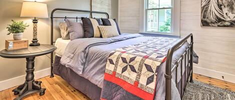 Un dormitorio con cama, una mesita de noche con lámpara, una ventana y una obra de arte colgada en la pared.