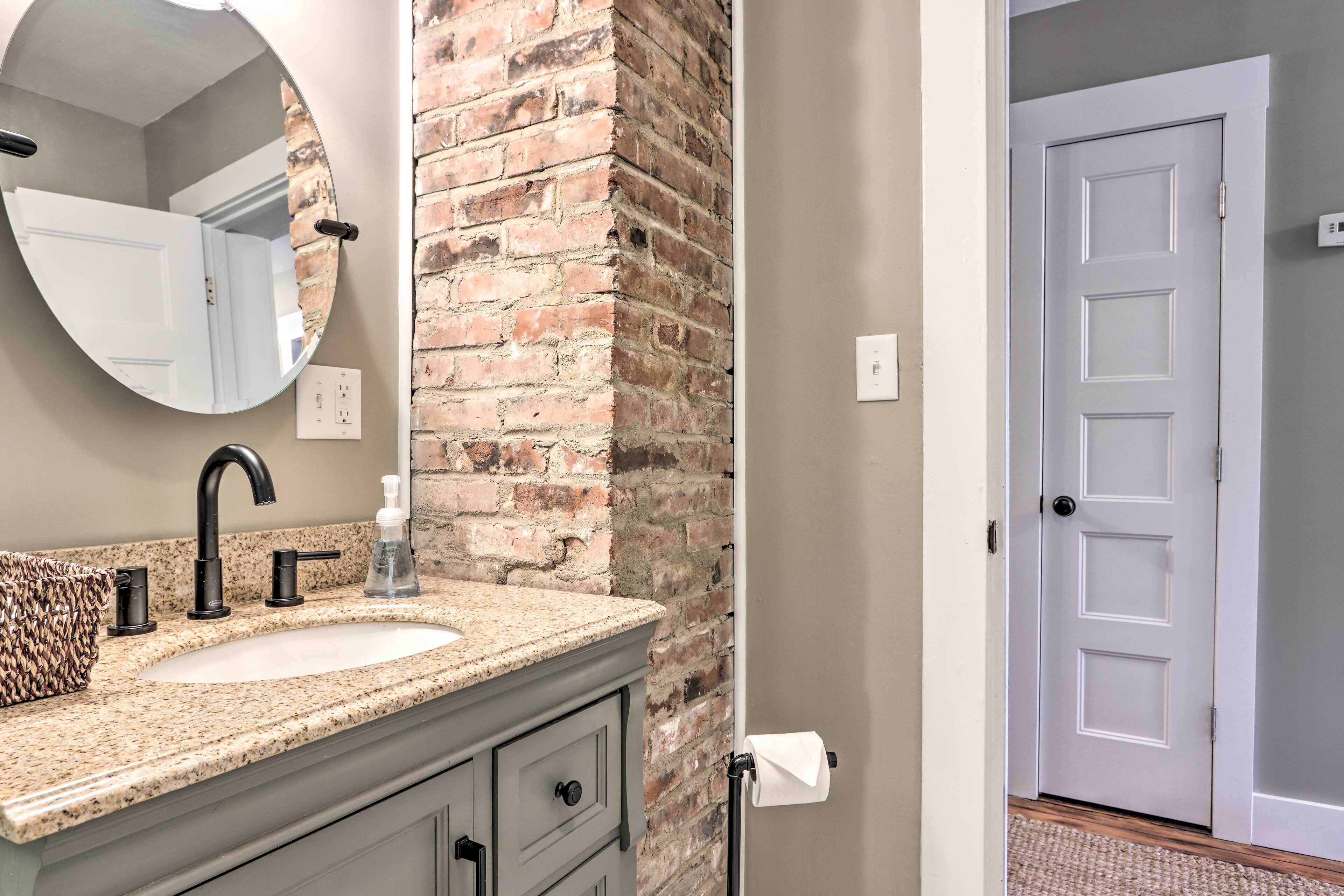 A bathroom with a granite countertop, a white sink, a round mirror, and a brick accent wall.