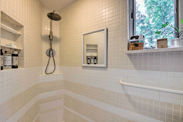 A bathroom with white tiled walls, a shower with a handheld showerhead, a window with a view of trees, and shelves with toiletries.