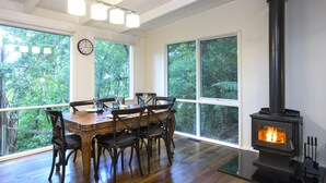 A dining area with a wooden table and chairs, a black wood-burning stove, and large windows offering a view of greenery.