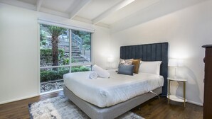 A bedroom with a large bed, a window with a view of a staircase and greenery, a bedside table with a lamp, and a wooden floor.