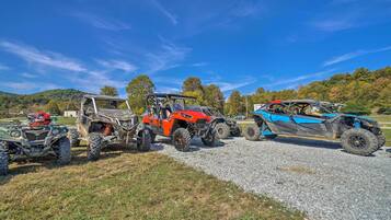A row of off-road vehicles parked on a gravel surface with a grassy area in the foreground and a forested hill in the background.