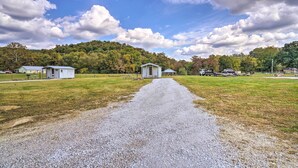 A gravel path leading to a small white building with a door, surrounded by a grassy field and a forested hill.