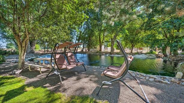 A serene outdoor seating area with a swing chair and a picnic table near a pond, surrounded by lush greenery. - Caldwell Country Home on Quiet Acreage (Caldwell)
