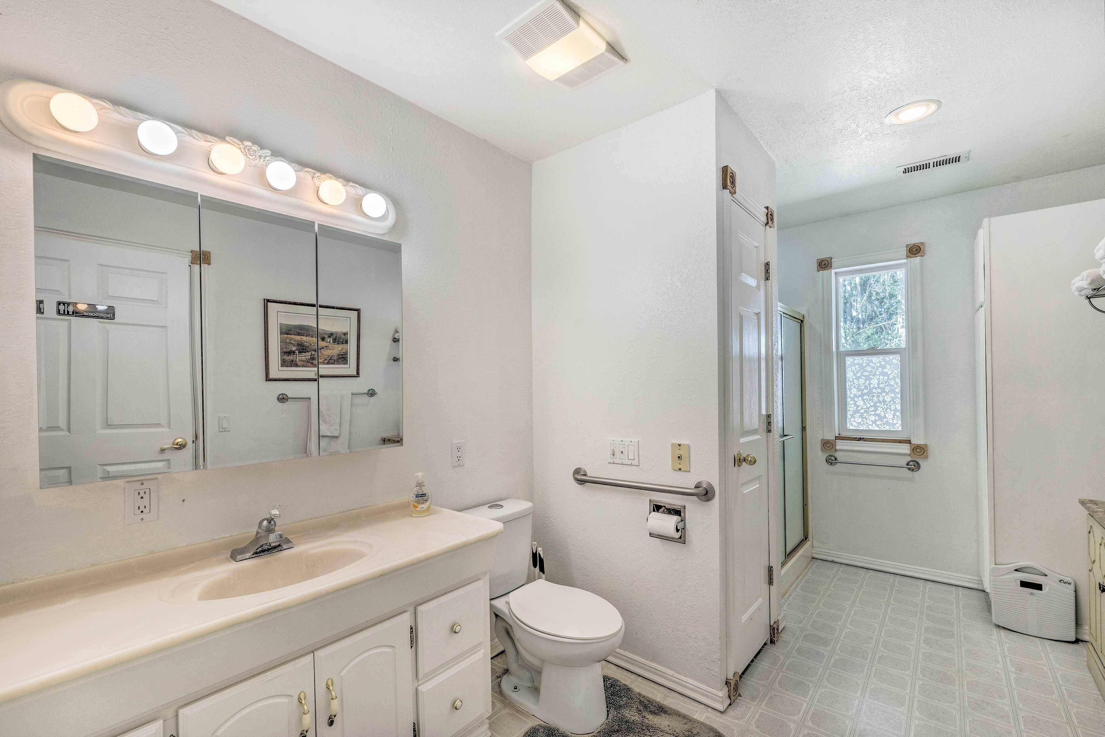 Une salle de bain équipée d’un meuble-lavabo, d’un évier, d’un miroir et de toilettes. Il y a une fenêtre donnant sur des arbres et une porte menant à une autre pièce.