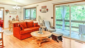 Un salon chaleureux avec une table basse en bois, un canapé rouge et un fauteuil gris. On y trouve également un coin repas avec une table et des chaises, ainsi que de grandes fenêtres donnant sur un paysage verdoyant.