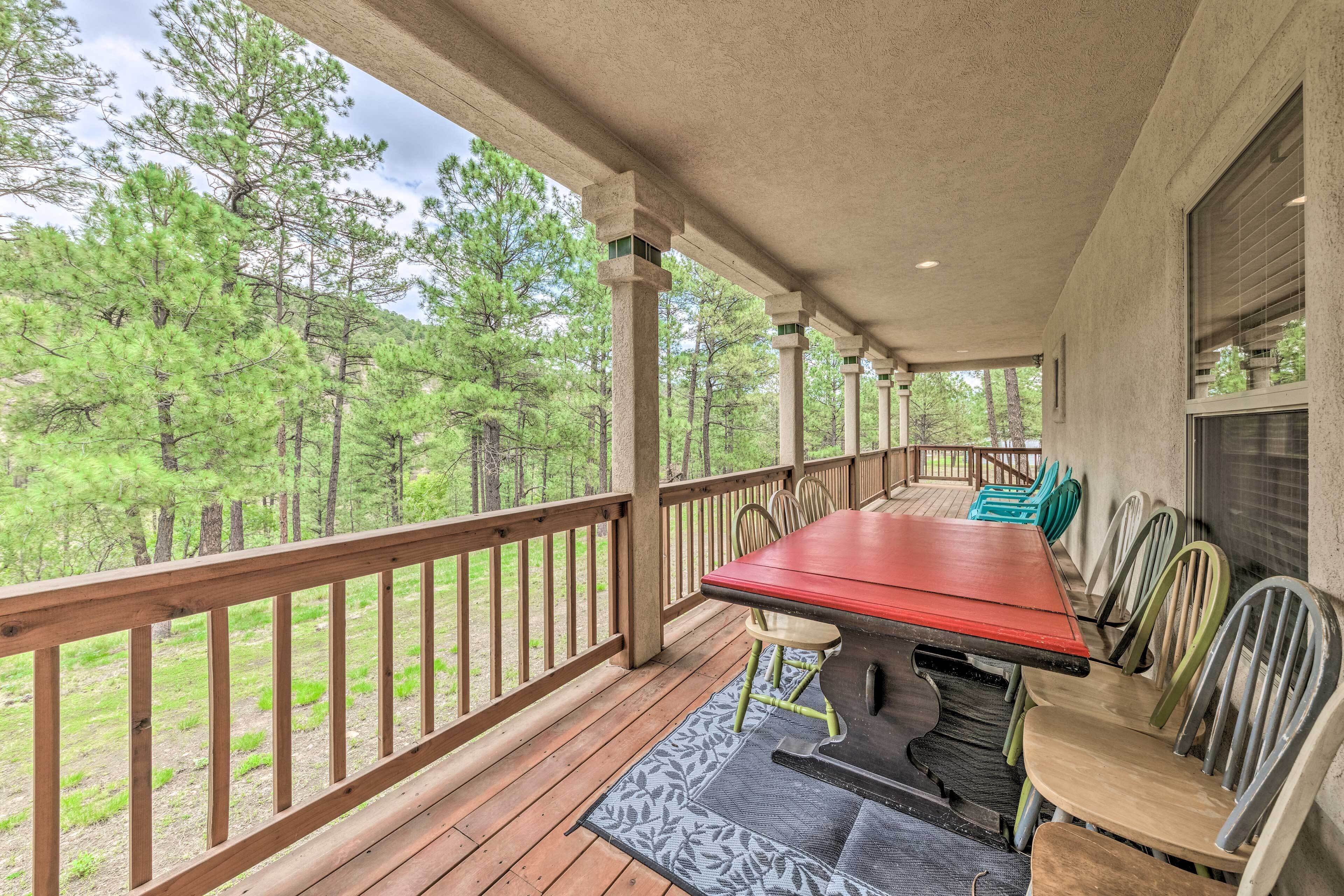 A covered outdoor deck with a wooden table and chairs, surrounded by a wooden railing, overlooking a forested area.