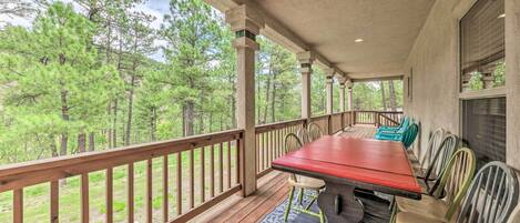 A covered outdoor deck with a wooden table and chairs, surrounded by a wooden railing, overlooking a forested area.