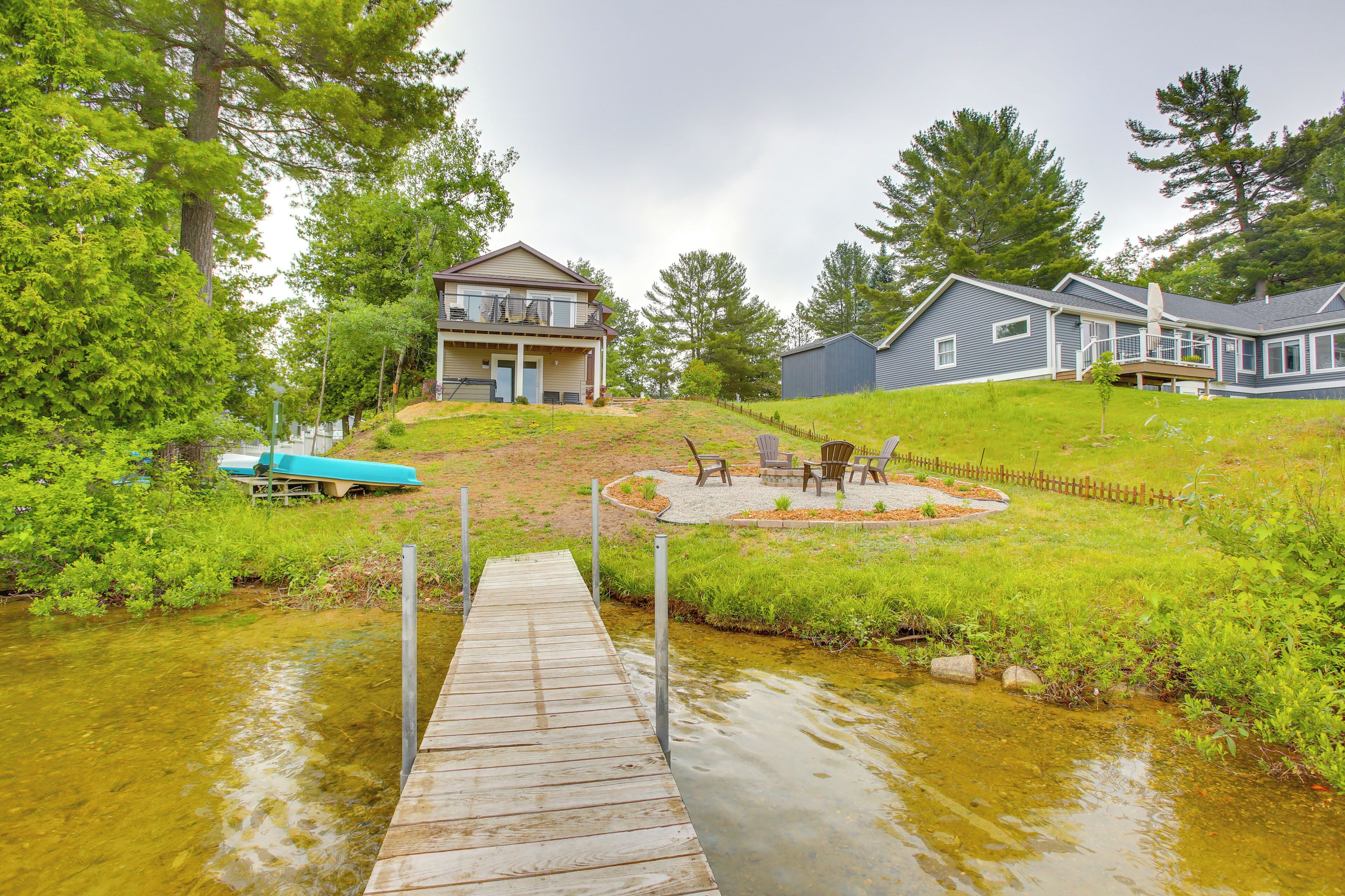 A residential area with a wooden dock leading to a house with a balcony, a backyard with a fire pit and chairs, and a small shed.