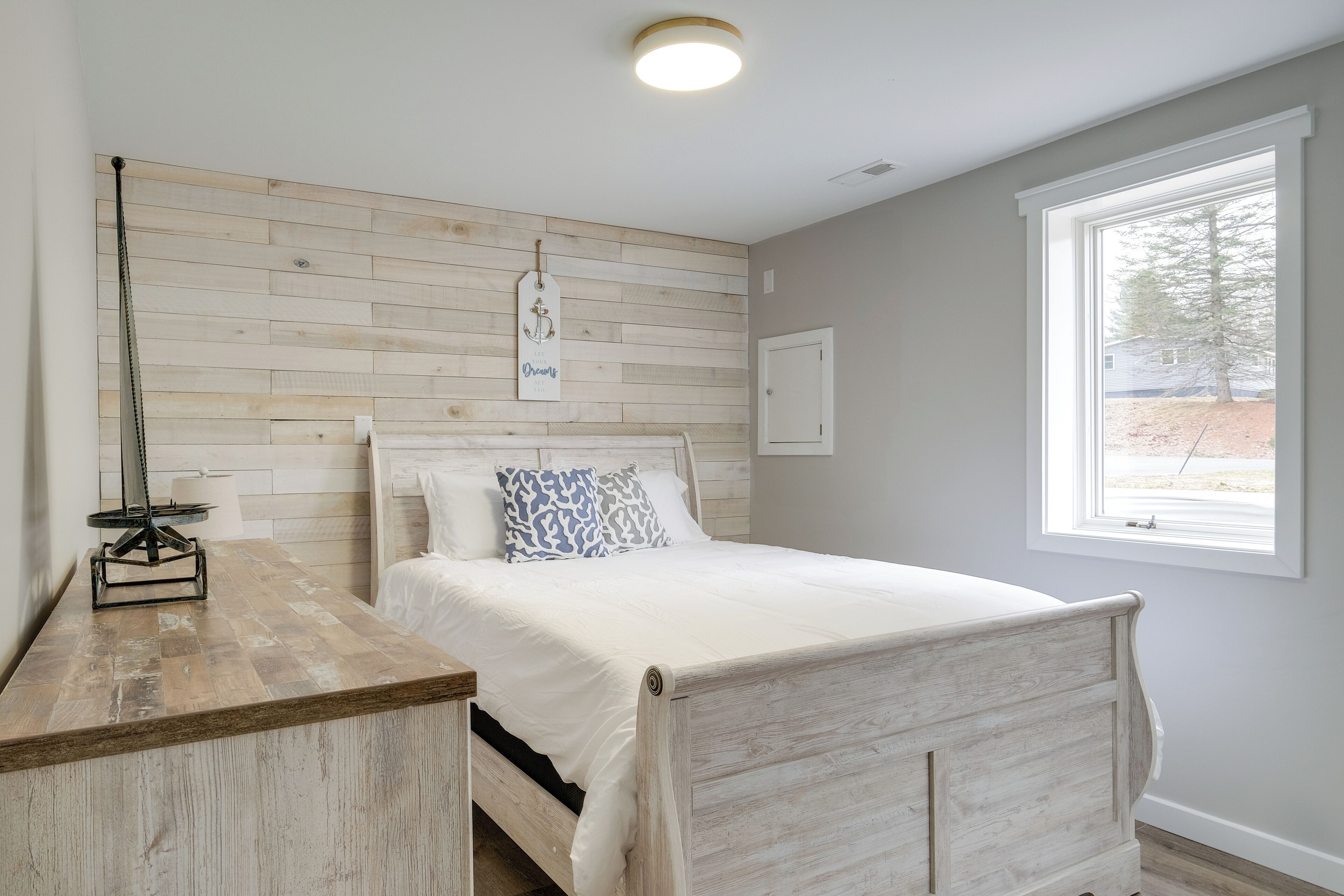 Een slaapkamer met een houten hoofdbord, een bed met wit beddengoed, een nachtkastje met een lamp, en een raam met uitzicht op het omliggende landschap.