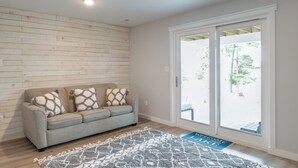 Ein Wohnzimmer mit grauem Sofa, gemusterten Zierkissen, einer Holzvertäfelung und einem großen Schiebeglasfenster zum Garten.