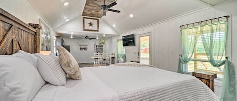 A bedroom with a large bed, wooden headboard, a ceiling fan, and a view of a kitchen area.