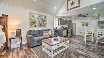 A spacious living room with a gray sofa, a wooden coffee table, and a kitchen area with cabinets and appliances.
