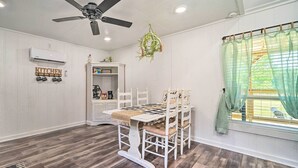 Un comedor con una mesa y sillas blancas, un ventilador de techo, un letrero de cocina y una ventana con cortinas translúcidas.