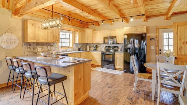 A spacious kitchen with wooden cabinets, a central island with bar stools, stainless steel appliances, and a hanging light fixture. - Modern Cabin w/ Grill Near Golf & Bass Fishing! (Burkesville)