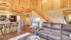 A spacious living area with a wooden staircase, a leather sofa, and a kitchen in the background. - Modern Cabin w/ Grill Near Golf & Bass Fishing! (Burkesville)