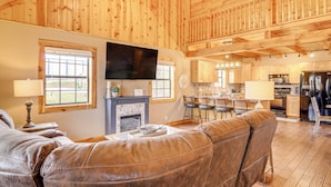 A spacious living room with a leather sofa, a fireplace, a flat-screen TV, and a kitchen area in the background. - Modern Cabin w/ Grill Near Golf & Bass Fishing! (Burkesville)