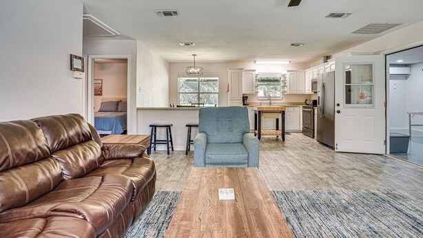 A spacious living room with a leather sofa, a blue armchair, and a wooden coffee table. A ceiling fan is visible, and the room has a large window providing natural light. - Granbury Cottage, Steps to Lake & Dock! (Granbury)