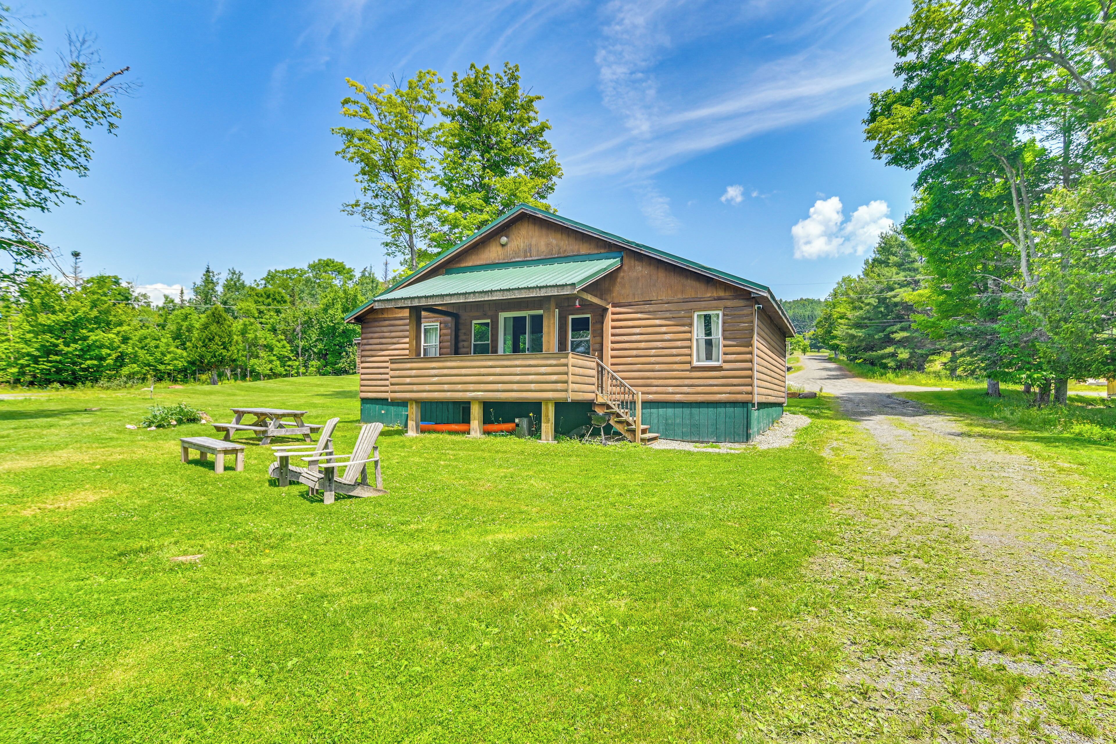 A wooden cabin with a green roof, surrounded by a grassy area with picnic tables and a dirt path.