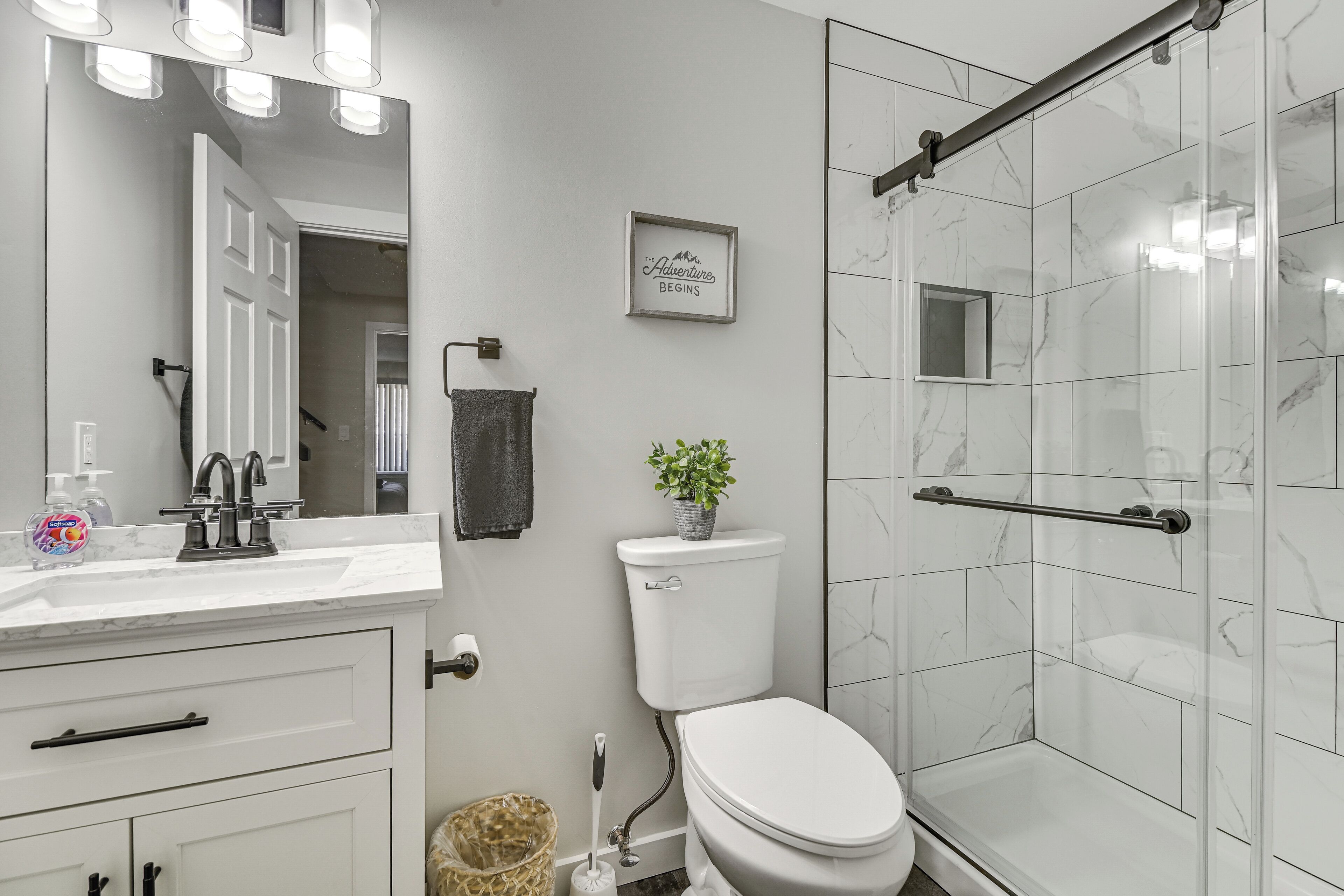 A bathroom with a white toilet, a sink with a mirror, a shower with a glass door, and a framed quote on the wall.