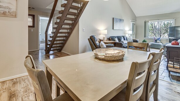 Un espace repas comprenant une table en bois, des chaises, un lustre, un escalier, ainsi qu’un salon avec un canapé et un téléviseur.