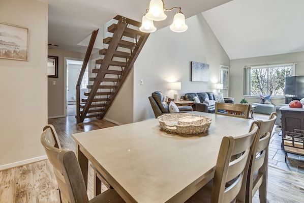 A dining area with a wooden table, chairs, a chandelier, a staircase, and a living room with a sofa and TV.