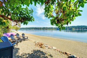A lakeside seating area with Adirondack chairs, a fire pit, and a view of the lake and forest. - Dreamy Wooded Cabin With Private Beach & Kayaks! (Olympia)