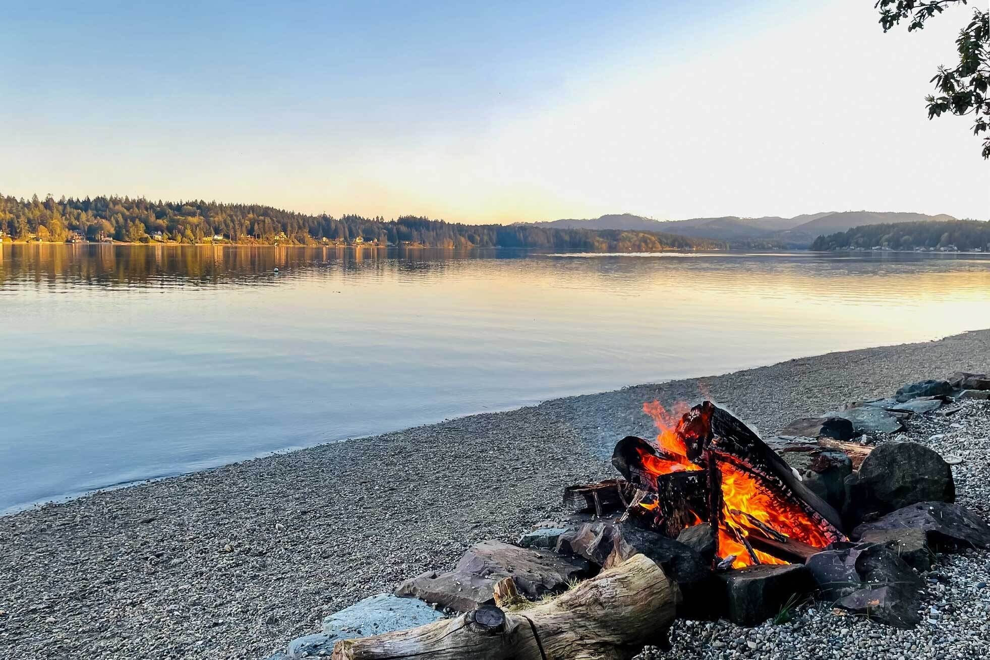 A lakeside view with a campfire on a pebbled shore.