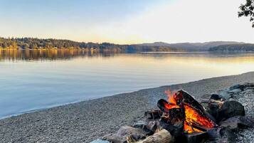 A lakeside view with a campfire on a pebbled shore.