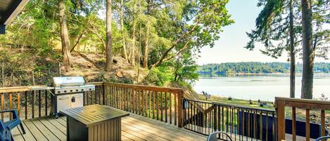 Terraza de madera con una parrilla, asientos y vistas a un lago rodeado de árboles.