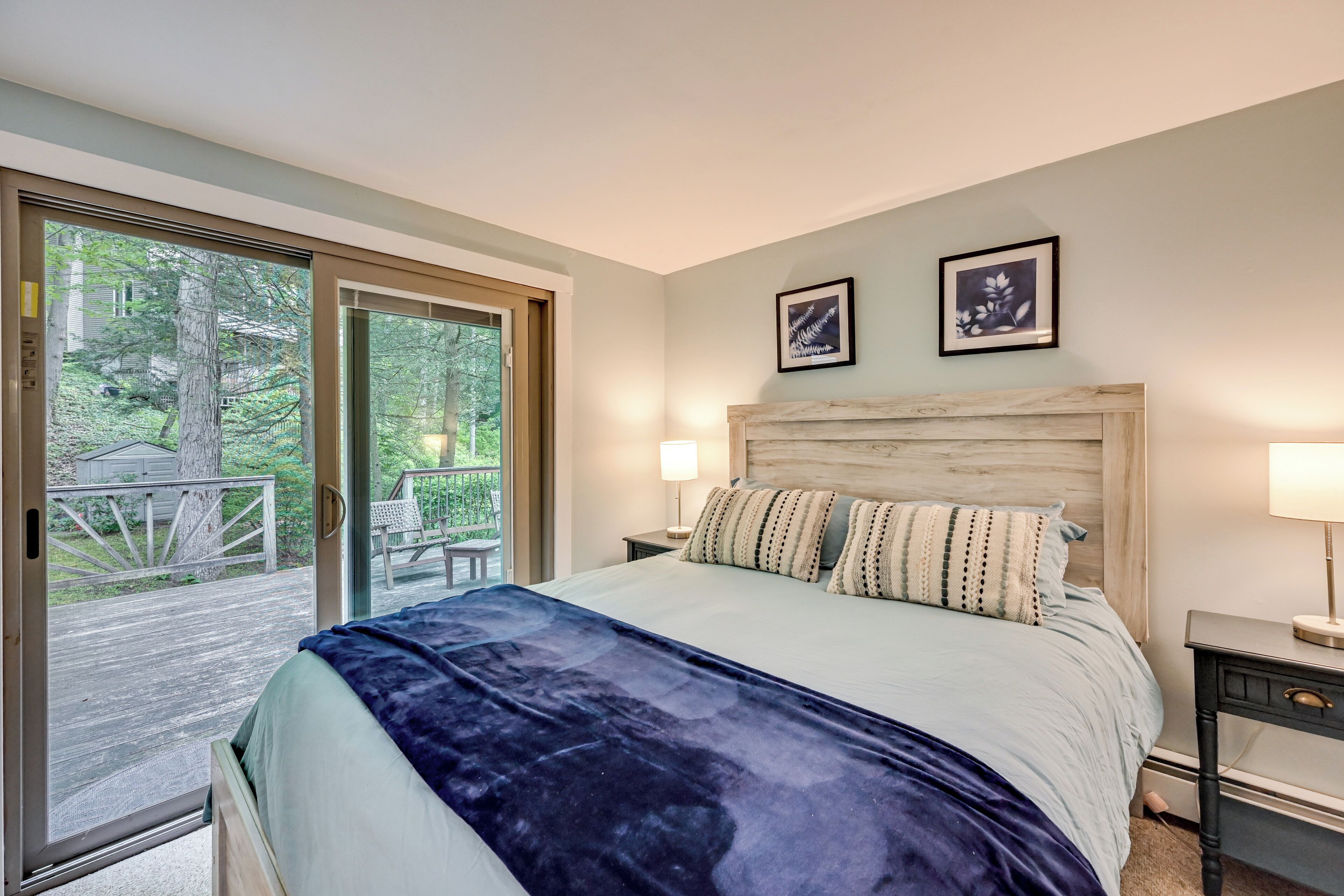Ein Schlafzimmer mit einem großen Bett, einem hölzernen Kopfteil, zwei gerahmten Bildern an der Wand und einer Glasschiebetür, die zu einer Außenterrasse führt.