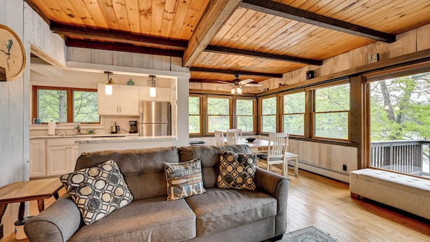 A spacious living area with a wooden ceiling, a gray sofa with patterned cushions, a dining table, and large windows offering a view of trees. - Lakefront Granby Escape w/ Private Dock & Kayaks! (Granby)