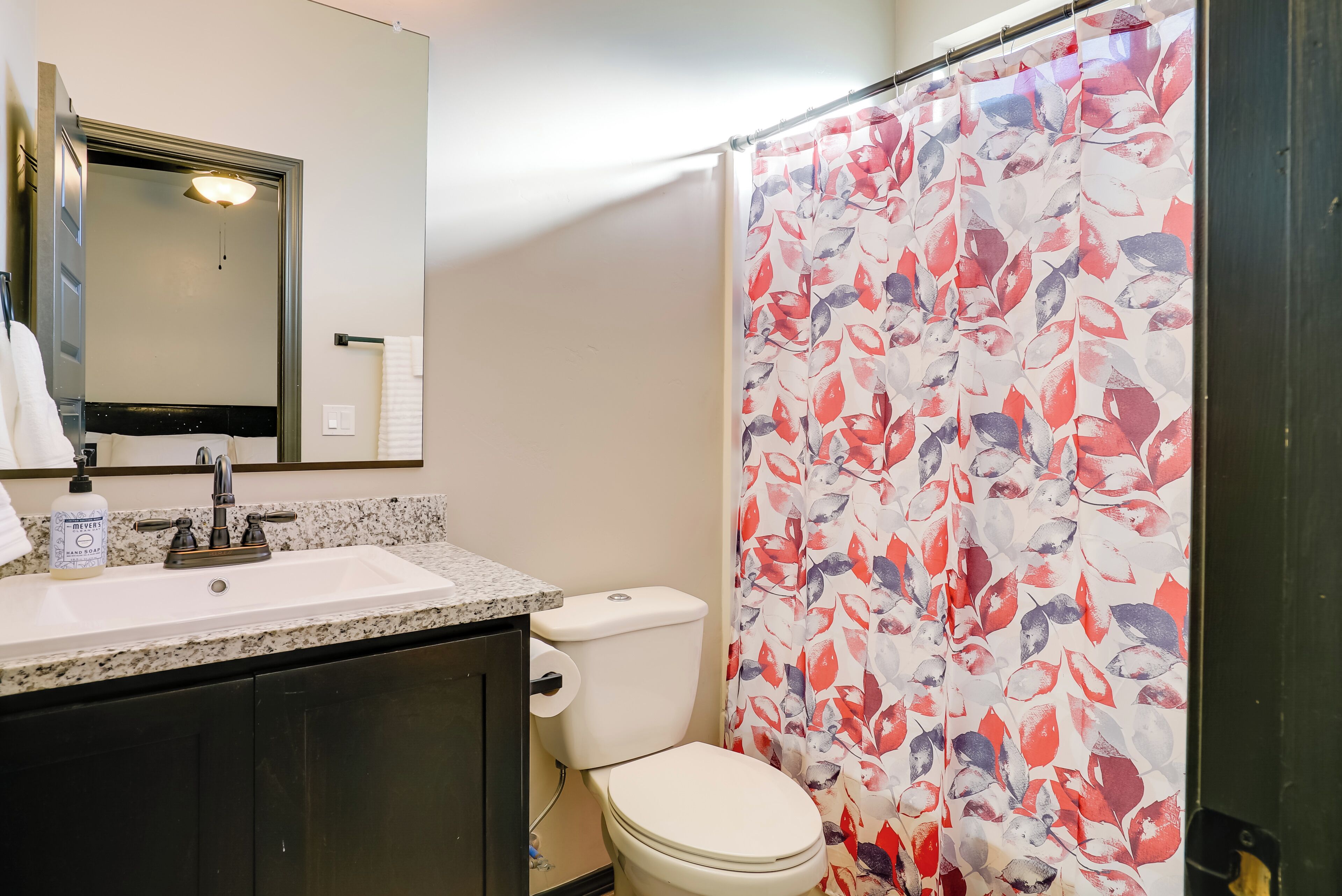 Une salle de bain avec un lavabo blanc, un miroir, des toilettes et un rideau de douche orné d’un motif de feuilles.
