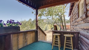 Une terrasse en bois avec un tapis vert, deux tabourets en bois et un bac à fleurs en bois orné de fleurs violettes.