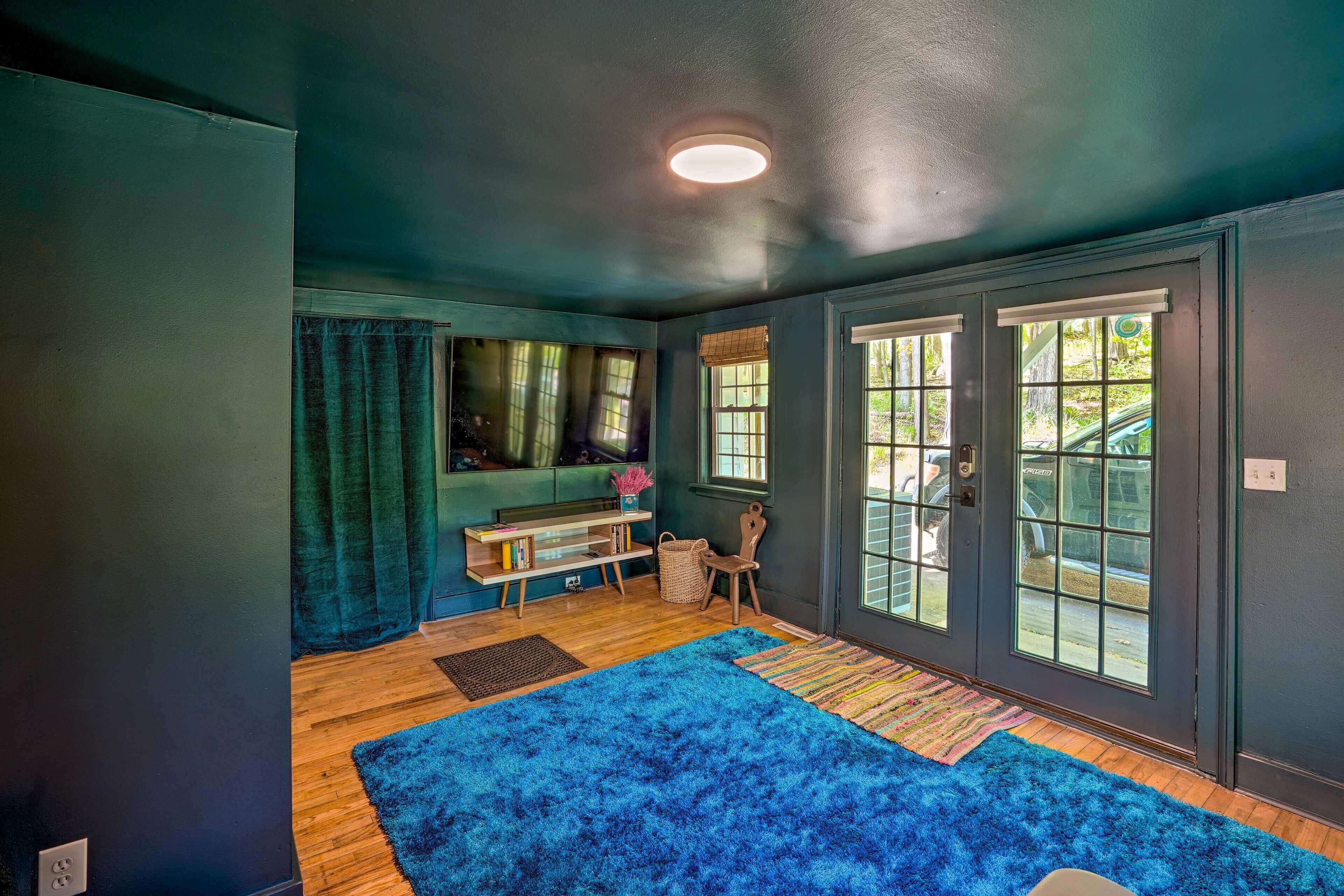 Una habitación con alfombra azul y mullida, suelo de madera, un escritorio con libros y una cesta, y ventanas amplias con persianas.