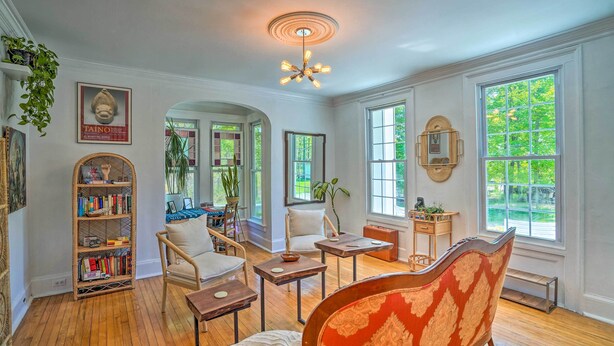 A spacious living room with hardwood floors, a large window with a view of greenery, a bookshelf, and a red patterned armchair. - Eclectic Troy Home w/ Hot Tub - Pet Friendly! (Troy)