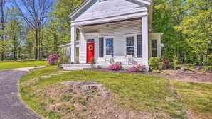 A white single-story house with a red door, surrounded by a well-maintained lawn and flowering plants. - Eclectic Troy Home w/ Hot Tub - Pet Friendly! (Troy)