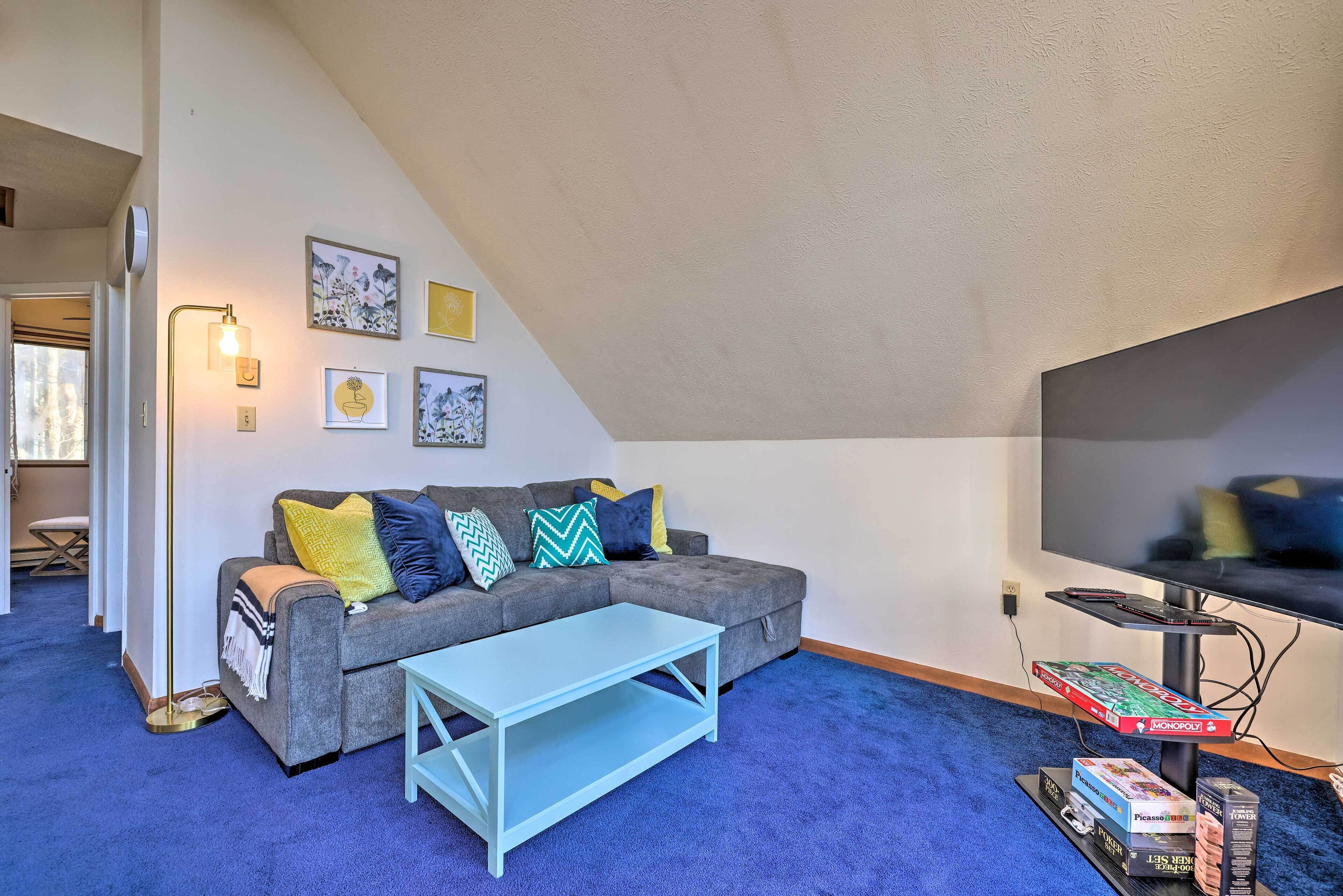 A living room with a grey sofa, a blue coffee table, and a flat-screen TV. There are framed pictures on the wall, a lamp, and a Monopoly board game.