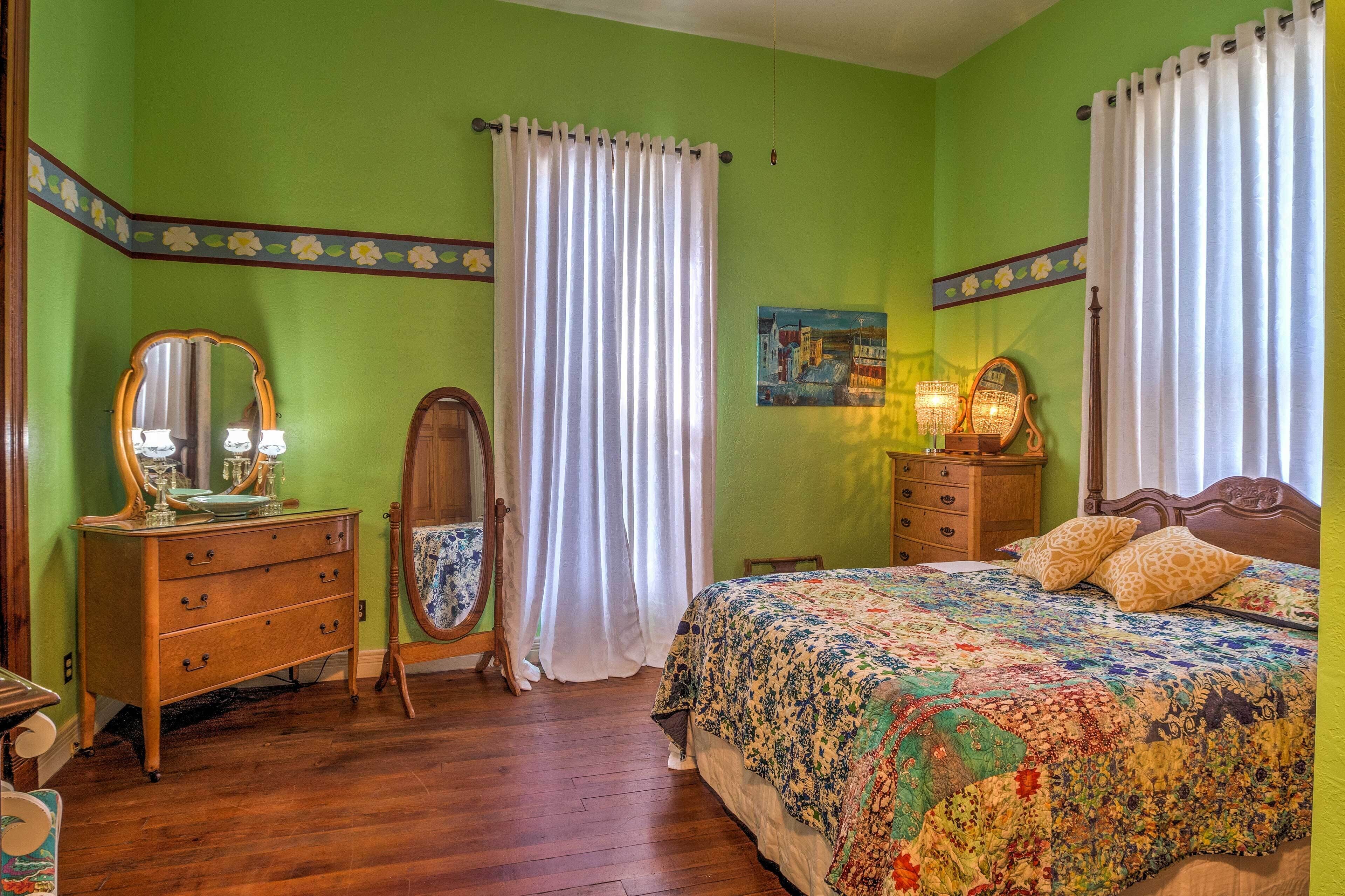 A bedroom with a wooden dresser, a bed with a floral quilt, a mirror, and a painting on the wall.