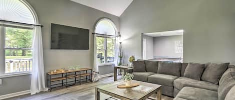 A spacious living room with a sectional sofa, a coffee table, and a TV mounted on the wall. There are large windows providing natural light and a view of the outdoors.