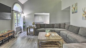 Ein modernes Wohnzimmer mit einem grauen Ecksofa, einem Holztisch und einer Bank. Ein Fenster mit weißen Vorhängen, ein Globus auf einem Beistelltisch und Kunstwerke an der Wand sind ebenfalls zu sehen.
