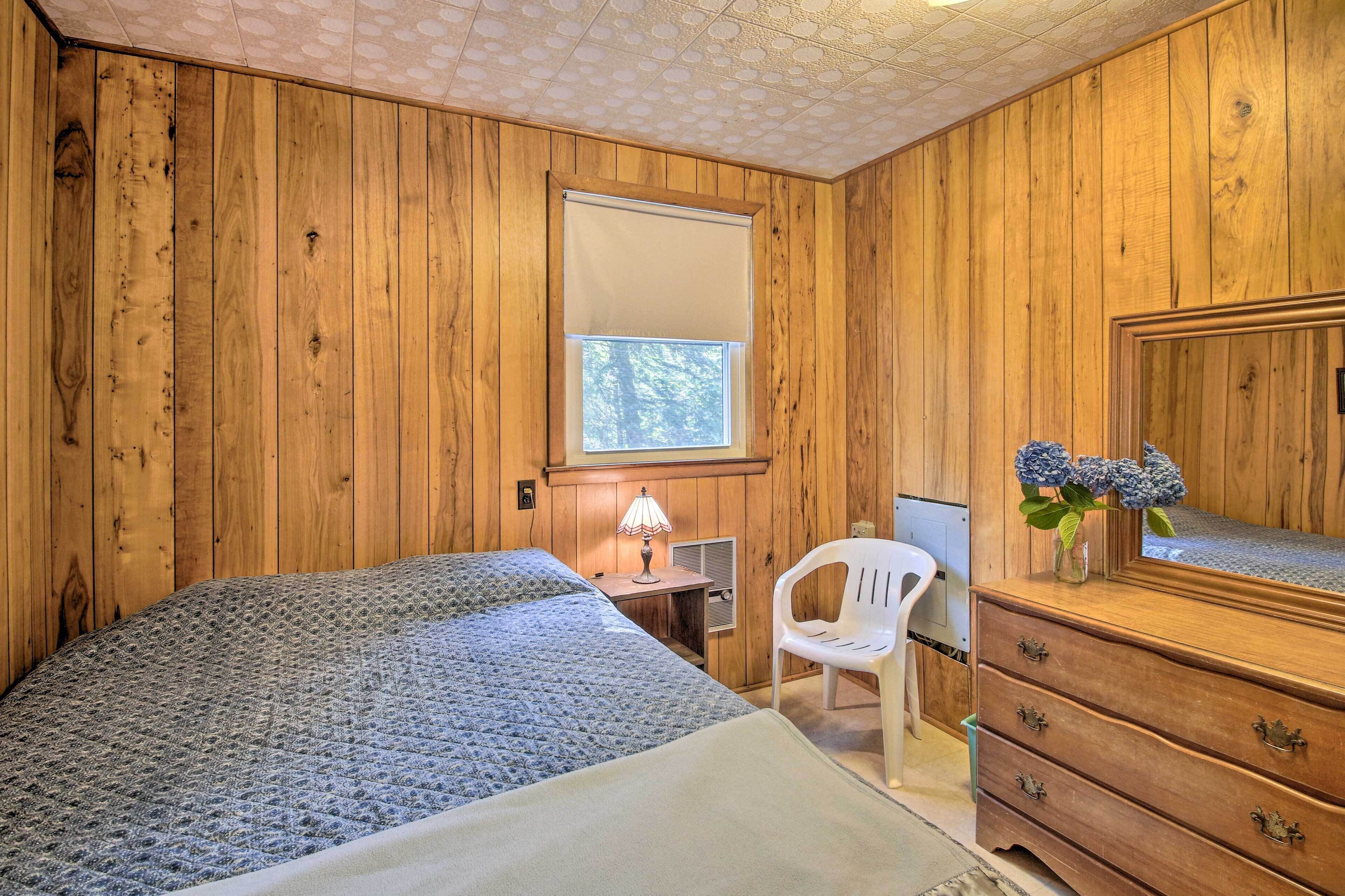 A wooden-paneled room with a bed, a dresser, a chair, and a window with blinds.
