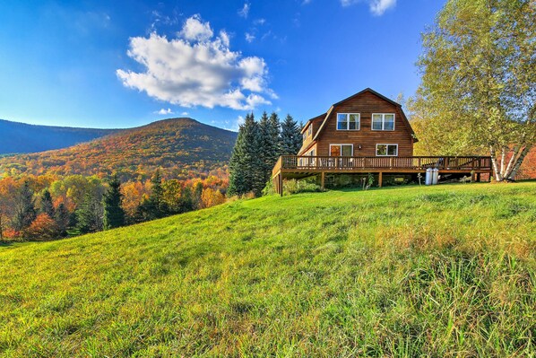 Eine Holzhütte mit einer Veranda, umgeben von einem grasbewachsen Hang und herbstlich gefärbten Bäumen, mit Blick auf ein Gebirge.