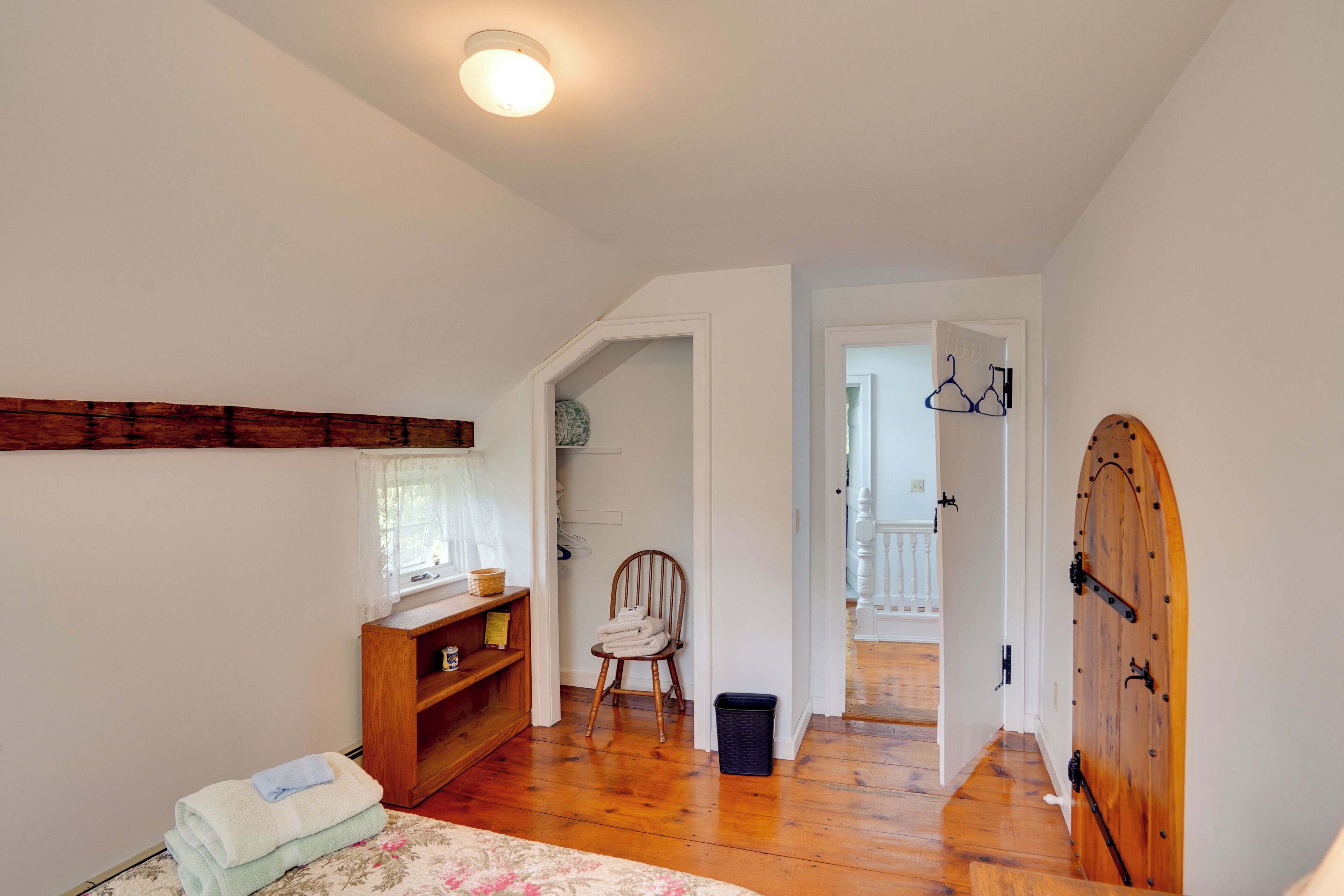 A room with wooden flooring, a chair, a small wooden shelf, a window with curtains, and a door with a decorative arch.
