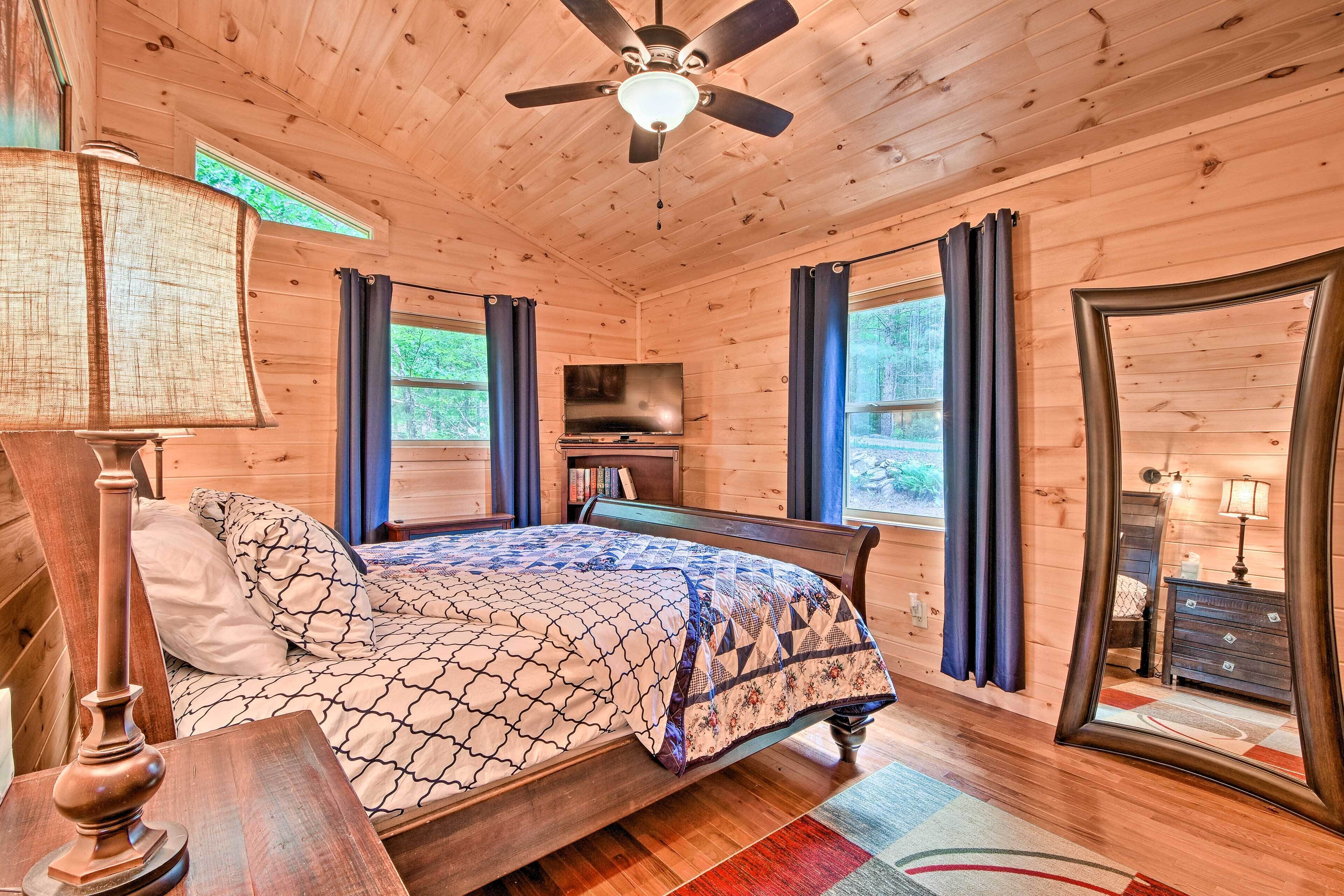 A wooden cabin bedroom with a bed, ceiling fan, TV, and large windows.