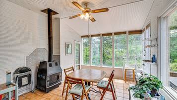 Quarto com mesa e cadeiras de madeira, ventilador de teto, lareira e amplas janelas com vista para o verde.